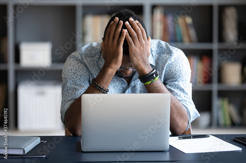 Unhappy hopeless African American man holding head in hands, overwhelmed tired businessman sitting at work desk with laptop, feeling exhausted, financial problem, loss money or bankruptcy