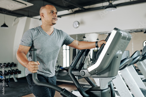 Serious mature mixed-race man working out on elliptical machine in gym to warm up before training