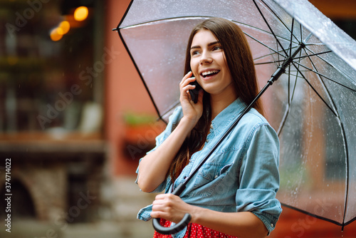 Smiling woman talking on mobile phone under umbrella. Young woman with a transparent umbrella at city street. Autumn concept.