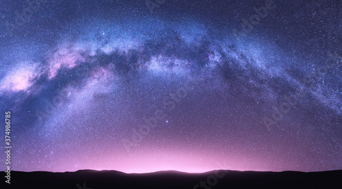 Milky Way arch. Fantastic night landscape with bright arched milky way, purple sky with stars, pink light and hills. Beautiful scene with universe. Space background with starry sky. Galaxy and nature