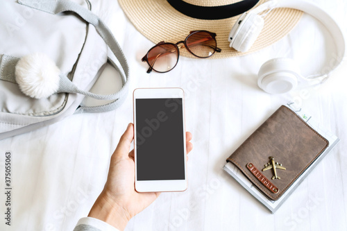 Woman hand holding smartphone with summer hat, sunglasses, bag, headphone, passport on bedroom. Prepare to travel, relaxation, journey, trip and vacation concepts. Top view and copy space.