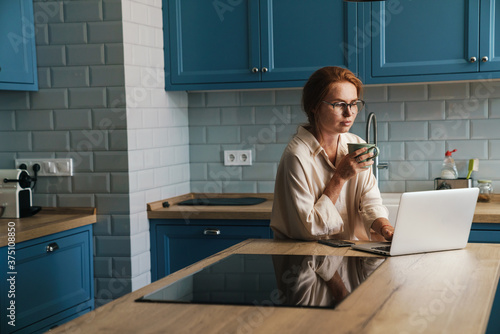 Image of drinking ginger woman drinking coffee and using laptop