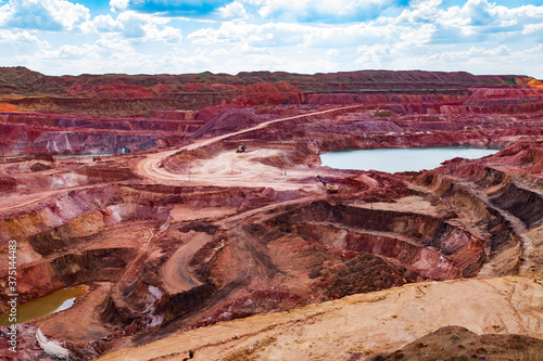 Aluminium ore quarry and blue lake in bauxite mine. Open cast (open-cut) mining. Excavators and quarry dump trucks. White road. On blue sky with clouds