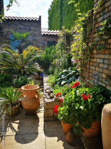 Widok na patio ze śródziemnomorską roślinnością. Akcenty w stylu prowansalskim. Ceglasty mur otoczony zielenią.