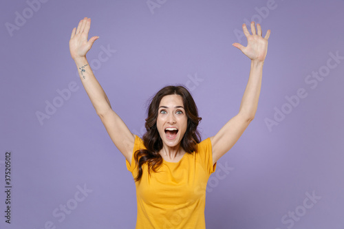 Excited surprised young brunette woman 20s wearing basic yellow t-shirt posing spreading rising hands up keeping mouth open looking camera isolated on pastel violet colour background, studio portrait.