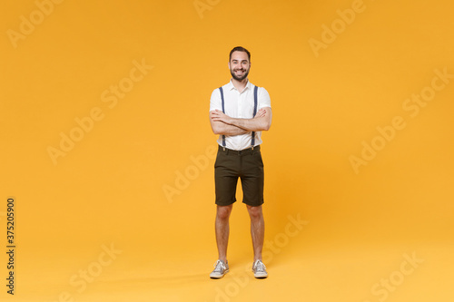 Full length portrait of smiling young bearded man 20s wearing white shirt suspender shorts posing standing holding hands crossed looking camera isolated on bright yellow color wall background studio.