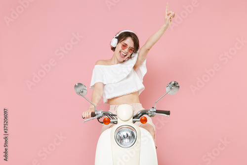 Cheerful young woman 20s in white summer clothes hat glasses listening music with headphones dancing pointing index finger up driving moped isolated on pastel pink colour background studio portrait.