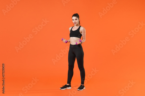 Full length portrait of beautiful young fitness sporty woman in black sportswear posing training working out doing exercise with dumbbells looking camera isolated on orange color background studio.