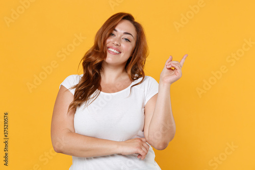Smiling young redhead plus size body positive female woman girl 20s in white casual t-shirt posing pointing index finger up on mock up copy space isolated on yellow color background studio portrait.