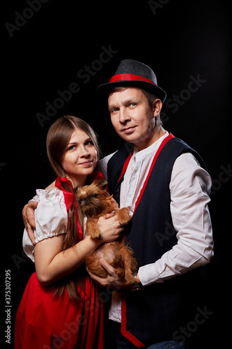 Couple in national ethnic clothing with small dog on hands posing in Studio on black background. Young woman and man or boy and girl in a red and black dress and pet Brussels Griffon in dark room.