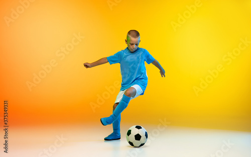 Kicking. Young boy as a soccer or football player in sportwear practicing on gradient yellow studio background in neon light. Fit playing boy in action, movement, motion at game. Copyspace.