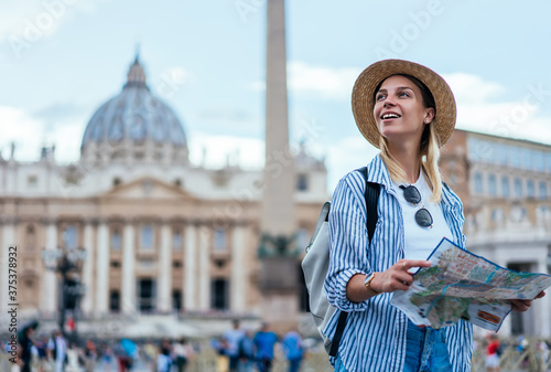 Smiling female tourist in straw hat enjoying beautiful architecture in Vatican feeling carefree during recreation journey, happy Caucasian hipster girl holding map having sightseeing tour near Rome