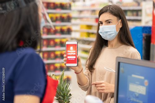 Modern woman wearing mask on face using digital discount coupon on her smartphone in supermarket