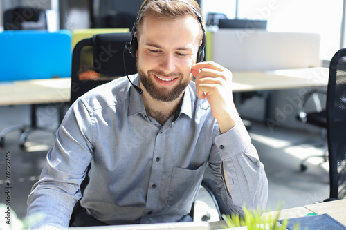 Smiling male call-center operator with headphones sitting at modern office, consulting online information in a laptop, looking up information in a file in order to be of assistance to the client.