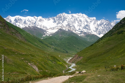 Beautiful mountain landscape in Georgia Svaneti. The snow-capped peaks of mount Shkhara near the Ushguli village are visible on a sunny day. A river flows between the green hills