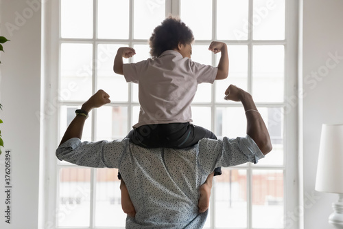 Back side view young african american father holding on shoulders little preschool funny kid son, showing biceps. Fit strong different generations family enjoying domestic activity, healthcare concept