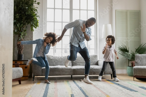 Happy african american father showing funny dancing moves to energetic small children siblings at home. Laughing crazy little biracial boy and girl having fun with caring daddy in living room.
