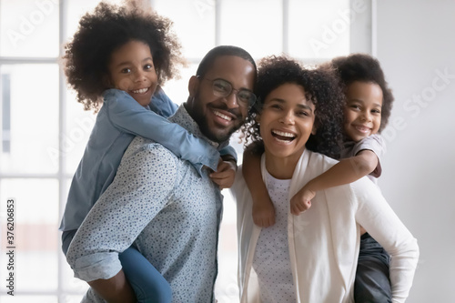 Portrait of happy biracial young couple parents giving piggyback ride to smiling adorable children siblings at home. Playful active family having fun with small kids indoors, looking at camera.