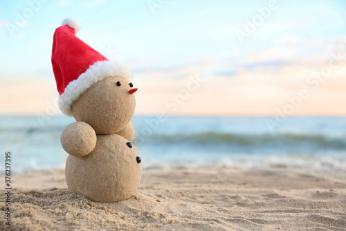 Snowman made of sand with Santa hat on beach near sea, space for text. Christmas vacation