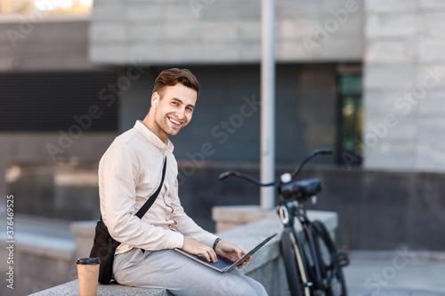 Networking outdoors. Happy guy in wireless headphones with coffee, works on laptop in city
