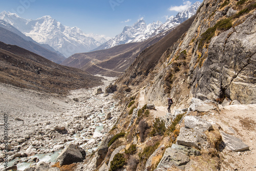 A female trekker is going down the valley in the Himalayas. Snow-capped peaks are in the background. Bright blue sky with a few white clouds.