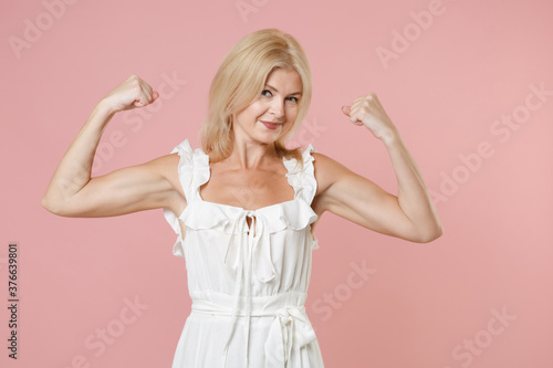 Smiling strong beautiful attractive young blonde woman wearing white summer dress spreading hands showing biceps muscles looking camera isolated on pastel pink colour background, studio portrait.