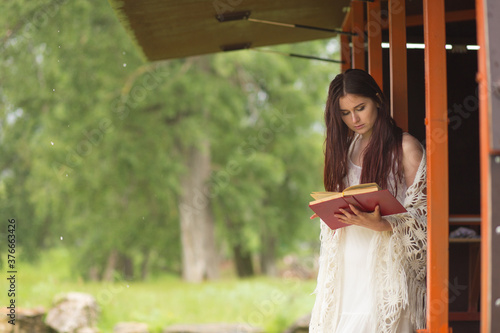 Pretty woman in white dress reading book in the cloudy nature. Girl sits on the terrace or on the porch