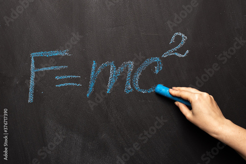 The physical formula of Einstein's theory is written in blue chalk on a blackboard. FMC2 is written by a science teacher or student in the classroom.
