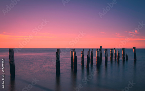 Tranquil seascape with ruined pilings of the pier in the seawater. Warm glowing pink sunrise over blue water. Long exposure image.