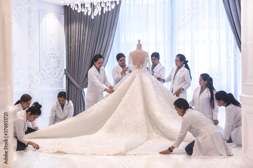Team of female dressmaking workers preparing a large haute couture wedding dress on a mannequin in a bridal design studio.