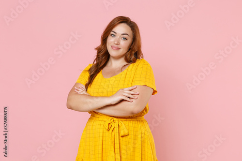 Smiling beautiful young redhead plus size body positive female woman girl 20s in yellow dress posing holding hands crossed looking camera isolated on pastel pink color wall background studio portrait.