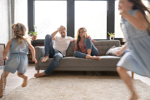 Tired mother and father sitting on couch feels annoyed exhausted while noisy daughters shouting run around sofa where parents resting. Too active hyperactive kids, need repose concept