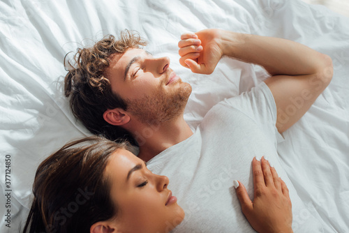 Top view of man stretching while lying on bed near sleeping girlfriend at morning