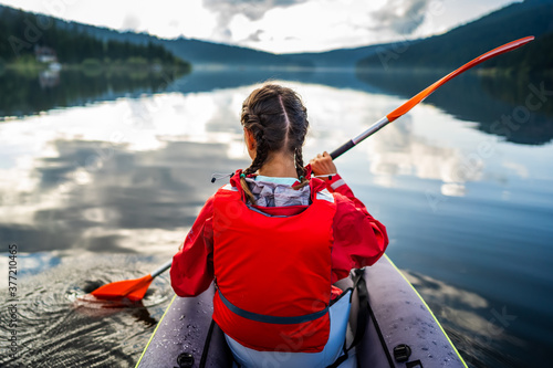 Caucasian woman in a kayak enjoys the clear view in front of her, with pine forest reflected in the beautiful and calm lake on a beautiful day of wild adventure. Hobby and leisure activity in nature.