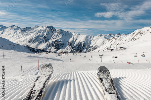 Panorama point of view skier legs on downhill start straight line rows freshly prepared groomed ski slope piste on bright day blue sky background. Snowcapped mountain landscape europe winter resort