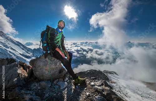 Climber in a safety harness, helmet, and high mountaineer boots with picturesque clouds background sitting at 3600m altitude on big rock and looking at summit during Mont Blanc ascending, France route