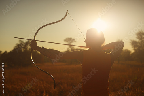 Young Caucasian female archer shooting with a bow in a field at sunset.