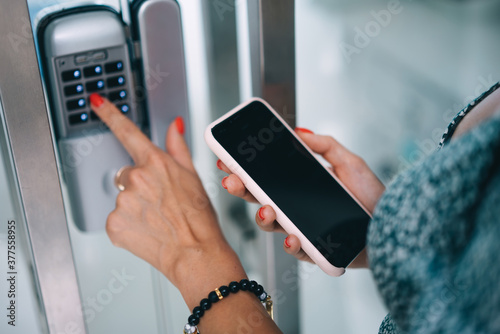 Cropped image of female's finder pressing buttons on electronic key panel checking code via smartphone modern application, woman unlocking door using gadgets to smart security home system