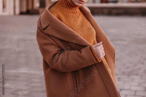 Young woman with cropped face in an orange knitted sweater and brown coat outdoor portrait in daylight. Cozy autumn fashion style. 