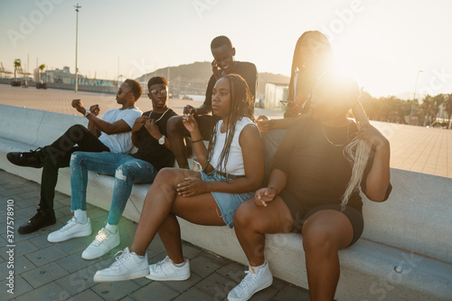 Group of young modern african black friends happily sitting together on the bench of the stairs in bright sunlight enjoying summer vacation