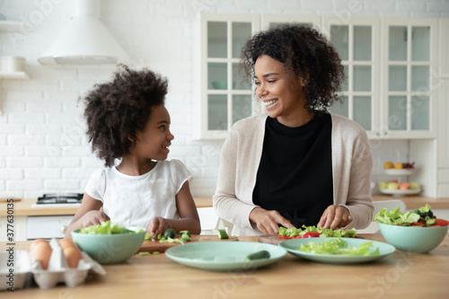 Happy small african american kid girl communicating with smiling biracial mother while preparing salad in modern kitchen, playful multiracial family having fun, enjoying cooking together at weekend.