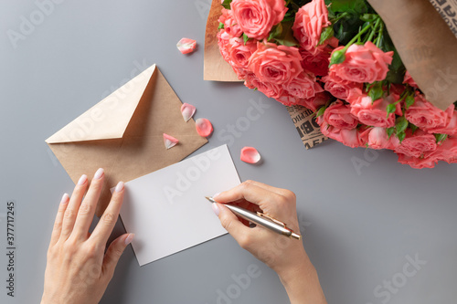 woman writing greeting card on gray table with bouquet of pink roses. Writing letter concept. top view.