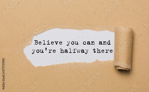 The text Believe you can and you're halfway there, appearing behind torn paper. Motivational quote. The craft paper is ripped.