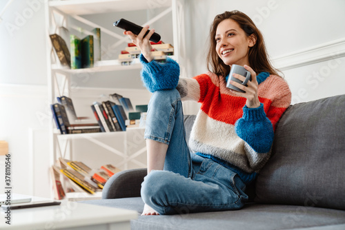 Photo of girl drinking coffee and using remote control while watching tv