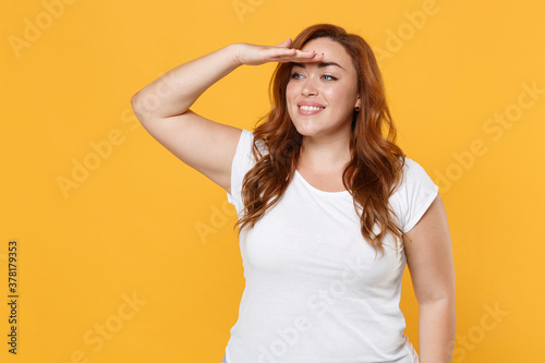 Smiling young redhead plus size body positive female woman girl in white blank casual t-shirt holding hand at forehead looking far away distance isolated on yellow color background studio portrait.