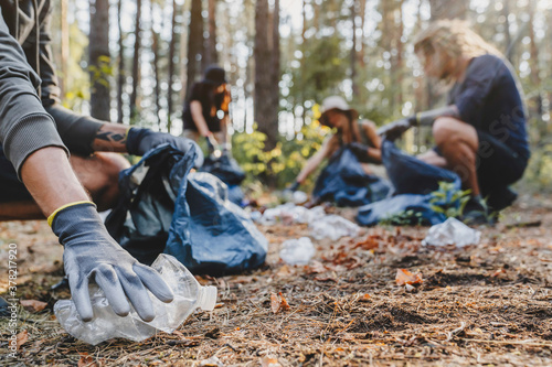 Close up of hands with plastic bottle with group of young people friends volunteers collecting plastic bottles to trash bags in forest background. Ecology concept