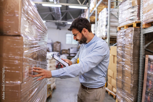 Supervisor in warehouse standing next to boxes and using tablet to check on goods.