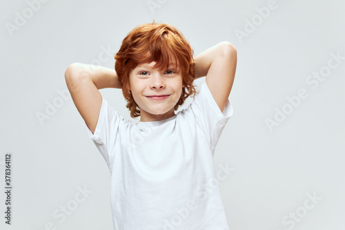 Cheerful red-haired boy holding hands behind his head smile white t-shirt 