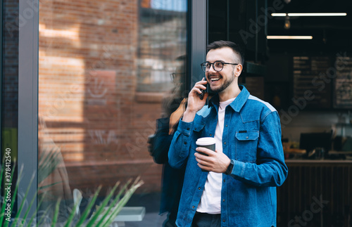 Cheerful male student talking on smartphone while standing with coffee near glass door of coffee shop
