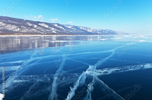 Landscape typical of winter Baikal Lake with beautiful blue smooth ice and snow-capped coastal mountains in February. Natural background. Winter ice travel on the lake (focus on ice)
